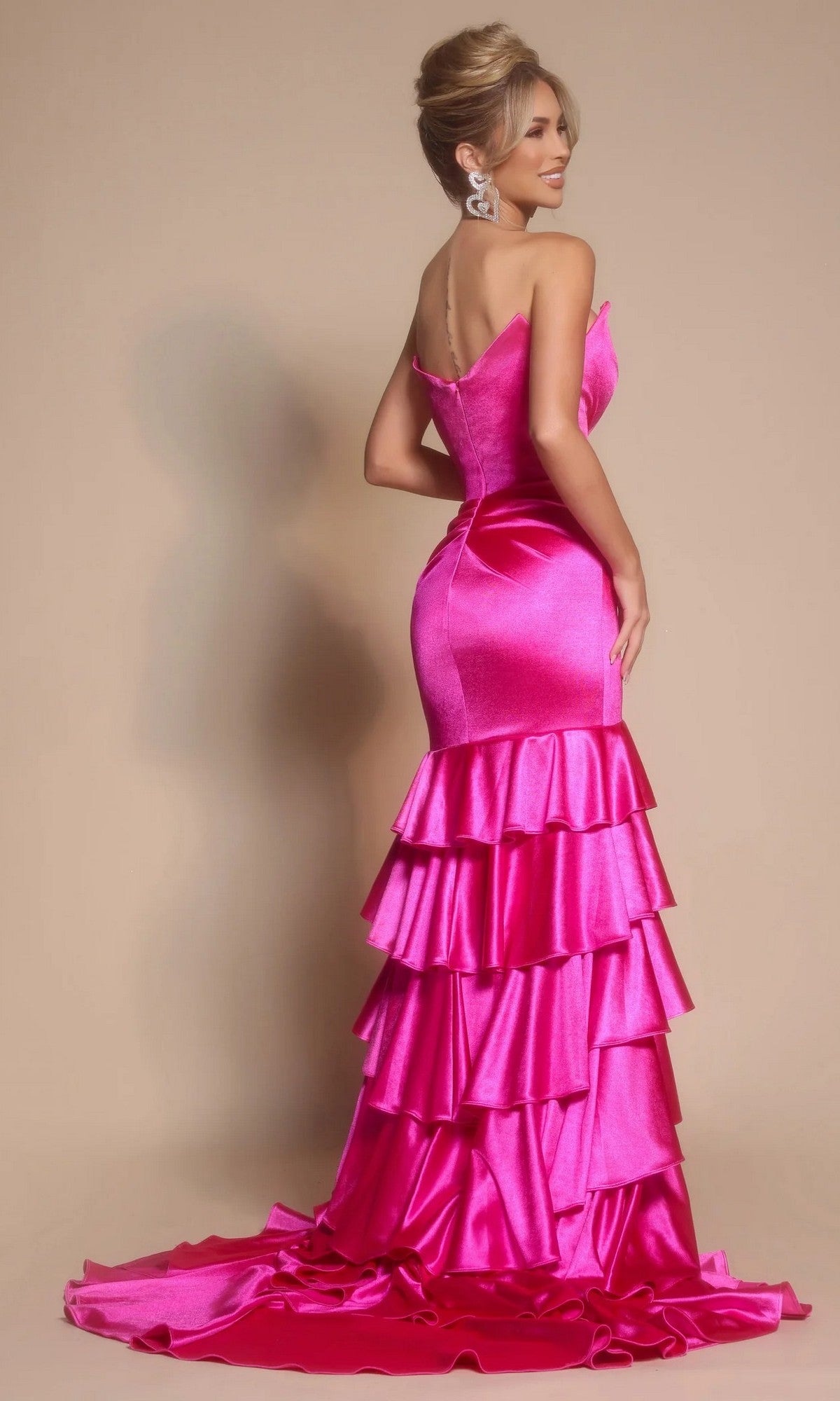 A woman poses elegantly with her back turned, wearing the Portia and Scarlett XO021 long prom dress—a strapless, bright pink satin gown with a fitted bodice and tiered ruffle skirt—against a beige background.